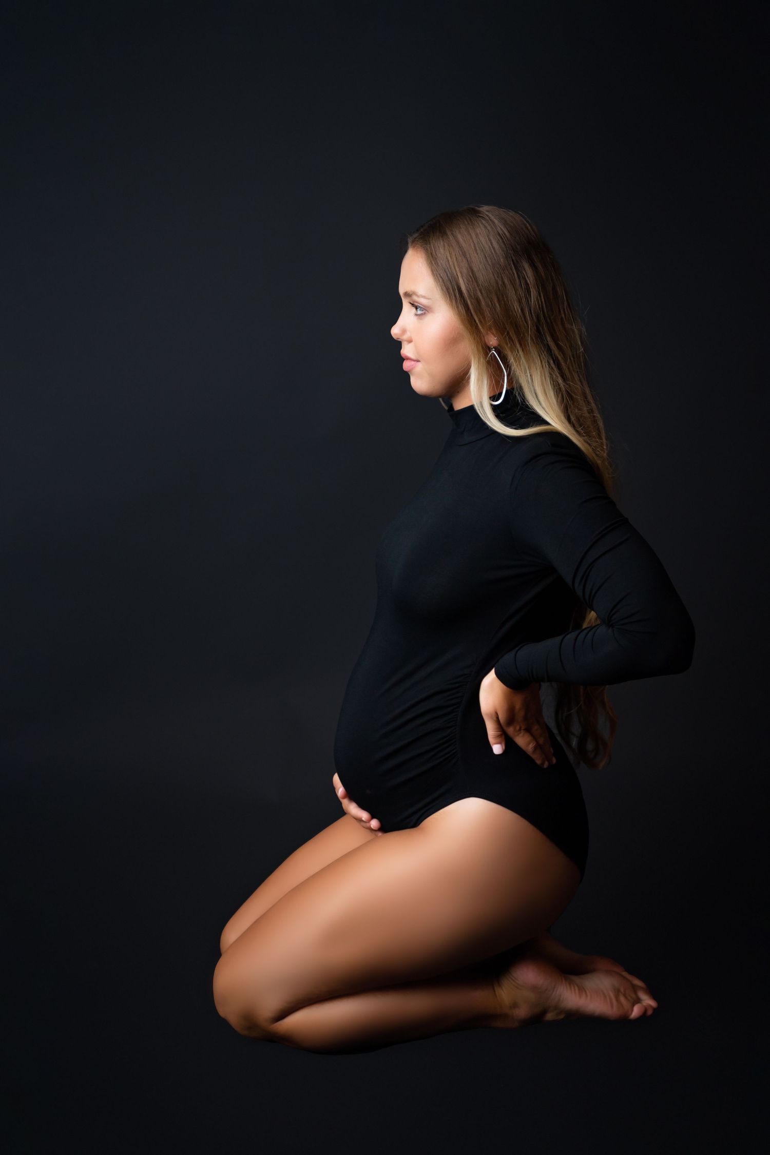 Pregnant woman in black bodysuit posing gracefully on knees against dark studio background.