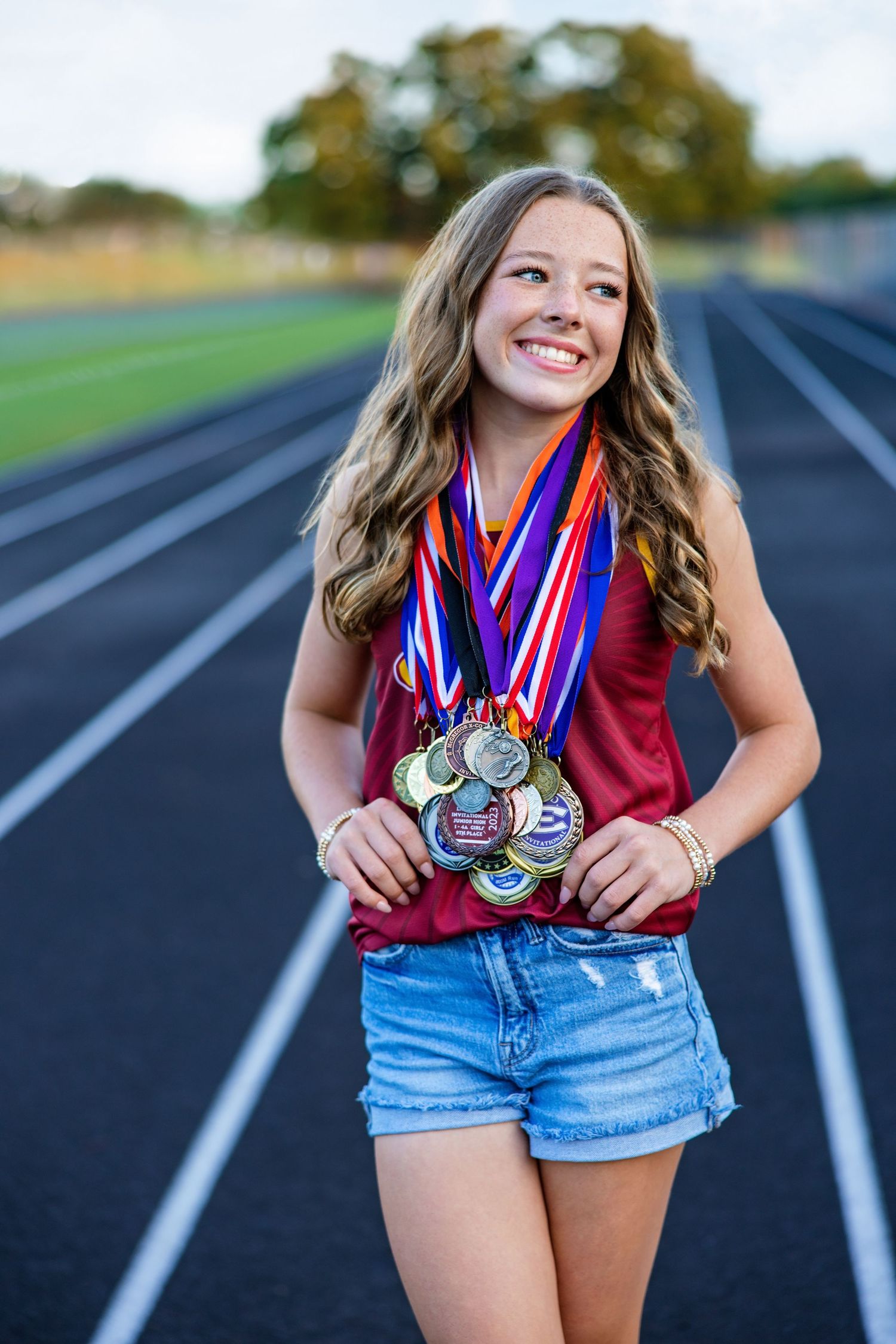 Teen girl smiling on track field wearing multiple sports medals and ribbons.