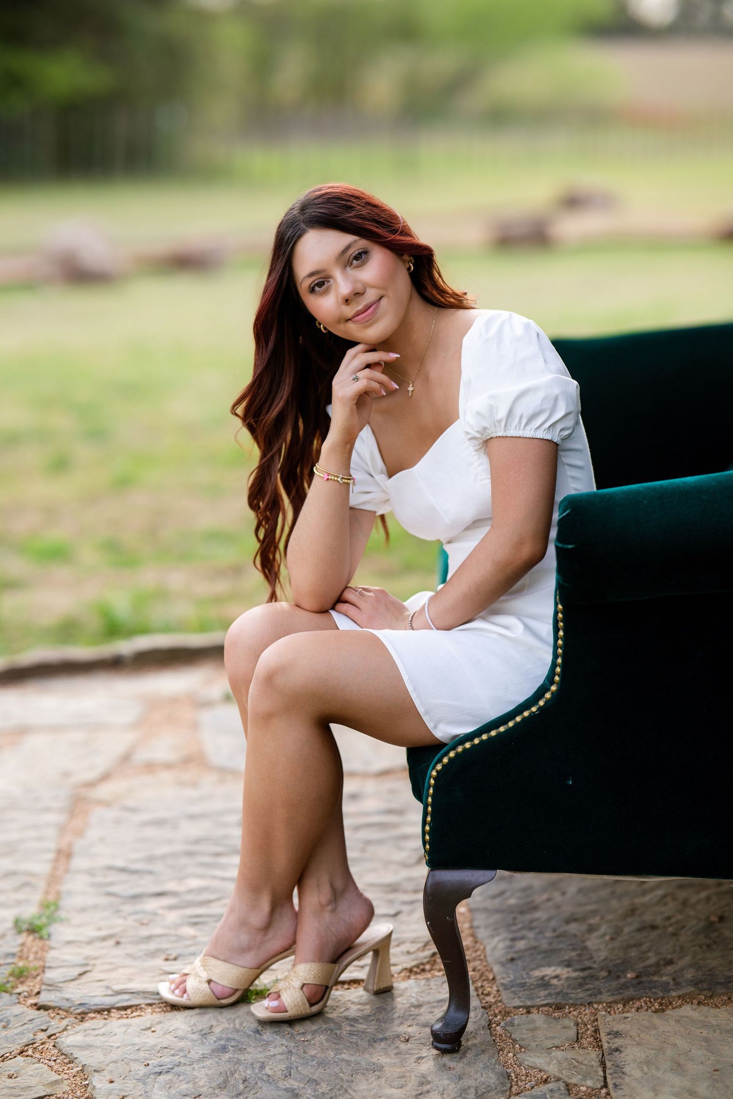 Brunette young woman in white mini dress sitting on green velvet chair outdoors for senior portraits.