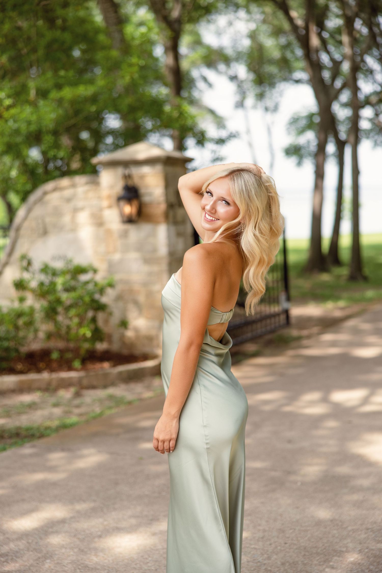 Blonde woman in elegant sage green backless satin gown posing outdoors near stone gate and trees.