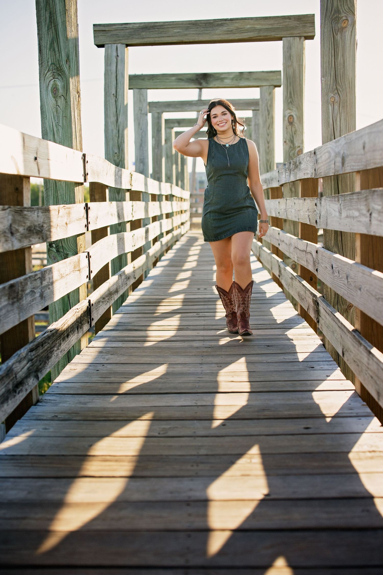 Woman in green dress and cowboy boots walks along rustic wooden boardwalk bridge.