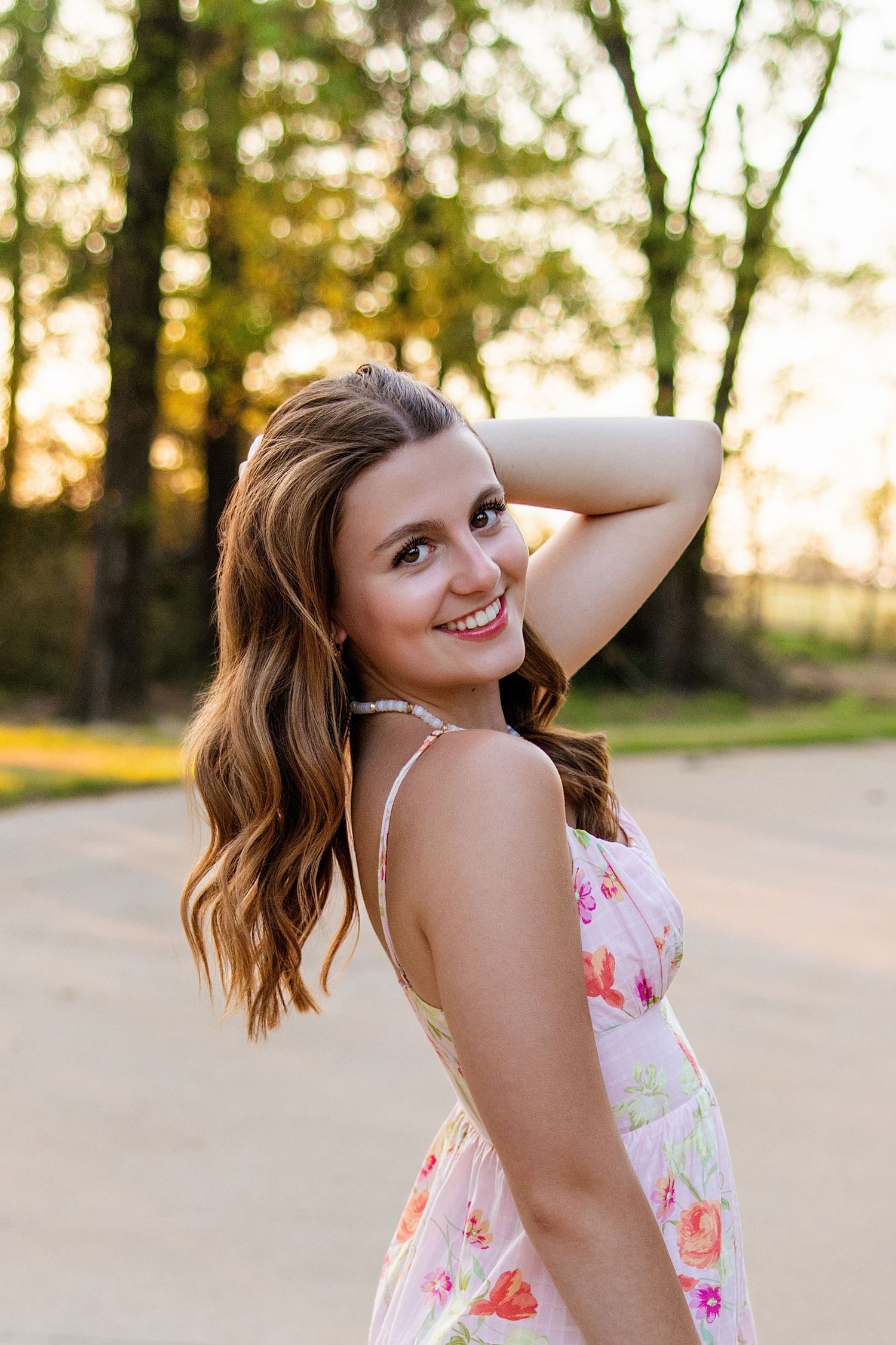 Smiling brunette teen girl in floral sundress posing outdoors at golden hour sunset for senior photos.