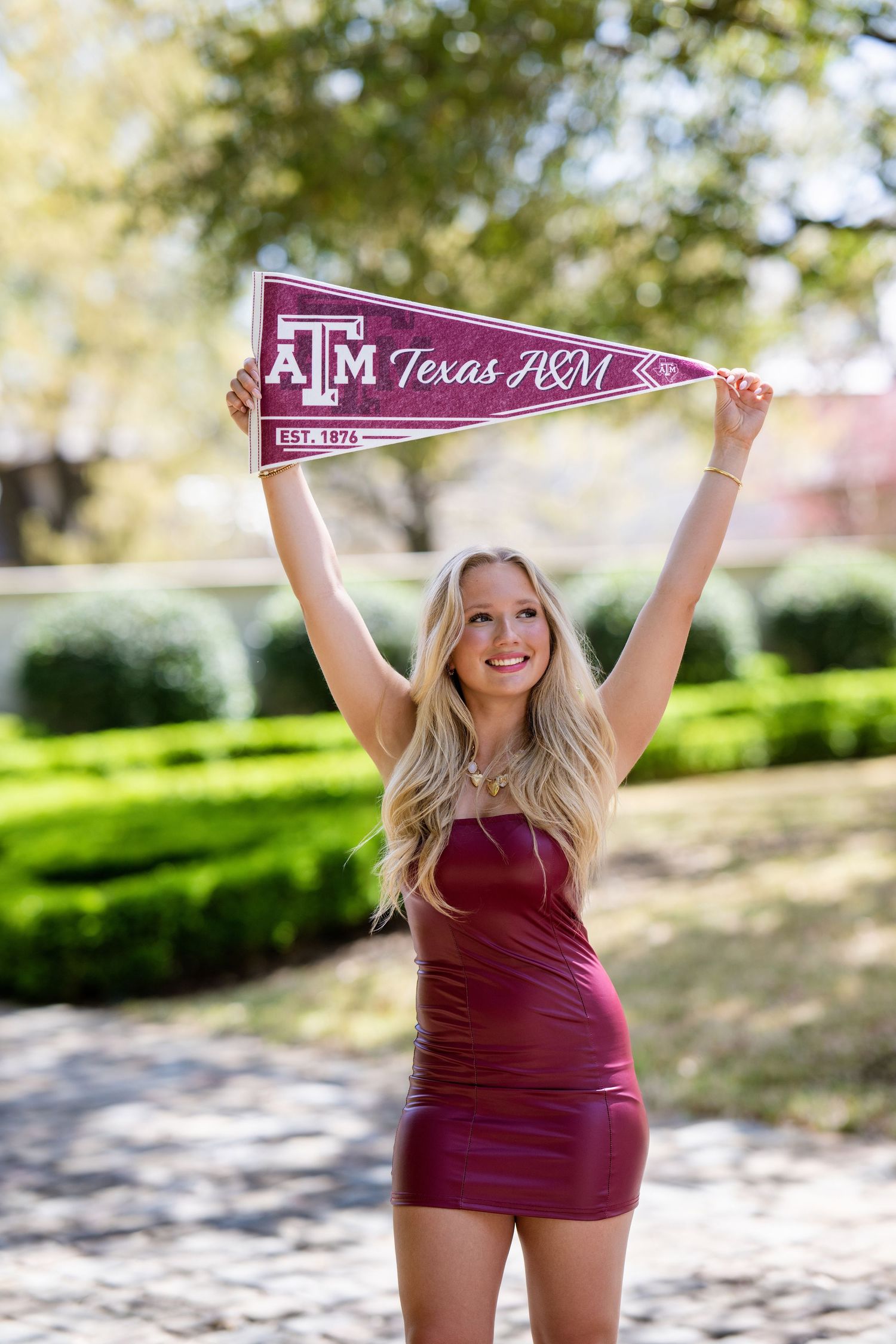 Smiling blonde woman in maroon dress holding Texas A&M pennant above her head outdoors.