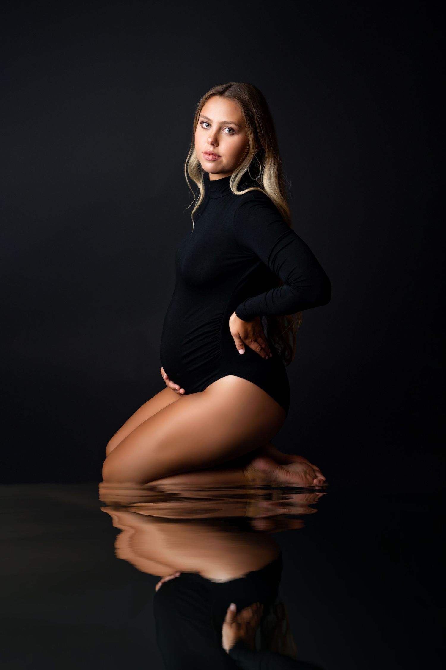Pregnant woman in black bodysuit kneeling on reflective floor against dark background.