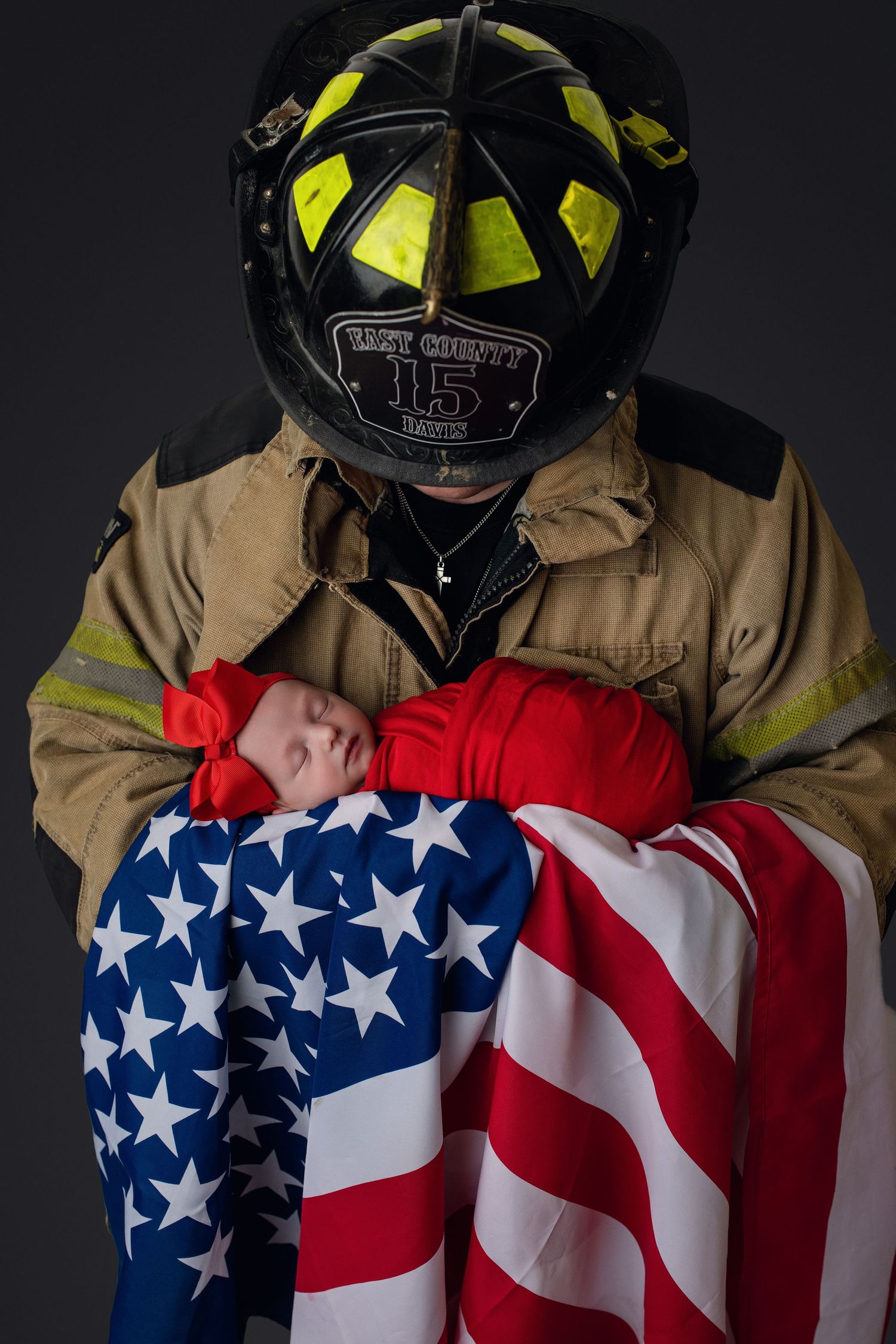 East County firefighter in full gear tenderly holds newborn baby wrapped in American flag.