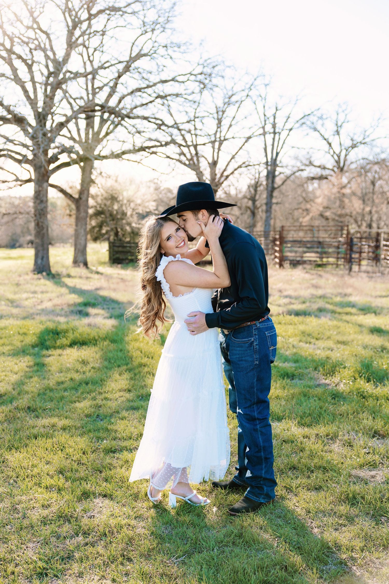 Romantic couple embracing outdoors, man in black cowboy hat kissing woman in white dress.