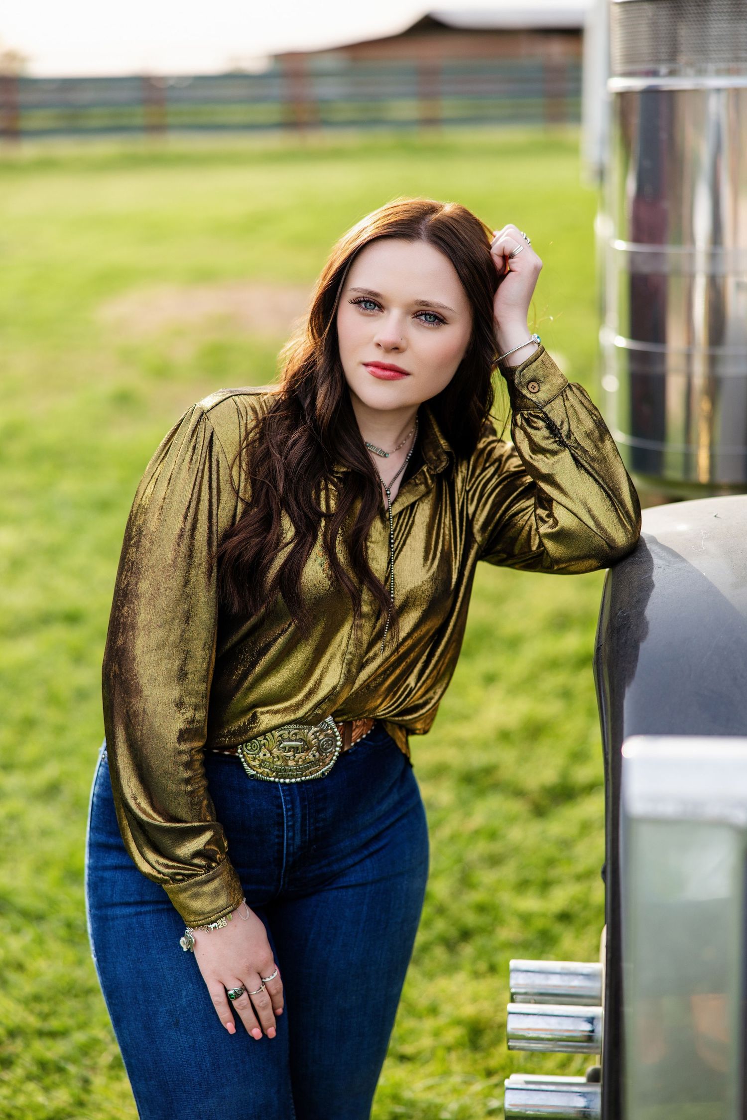 Young woman in gold metallic shirt and jeans leans against silver trailer on a farm at dusk.