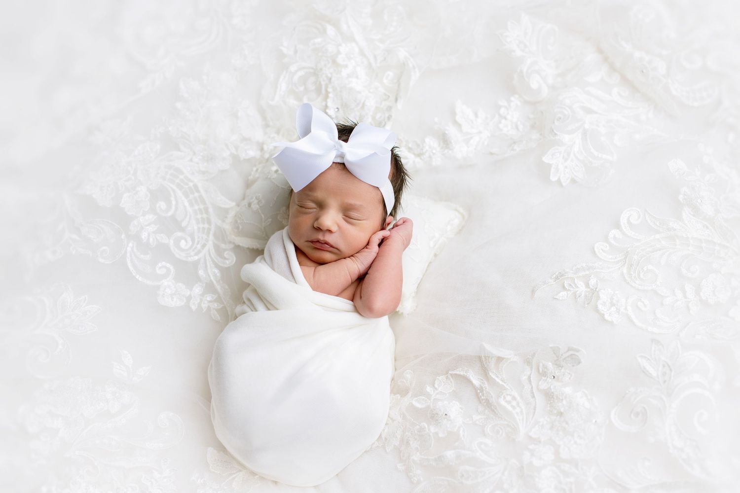 Sleeping newborn baby wrapped in white swaddle with bow headband on lace backdrop.