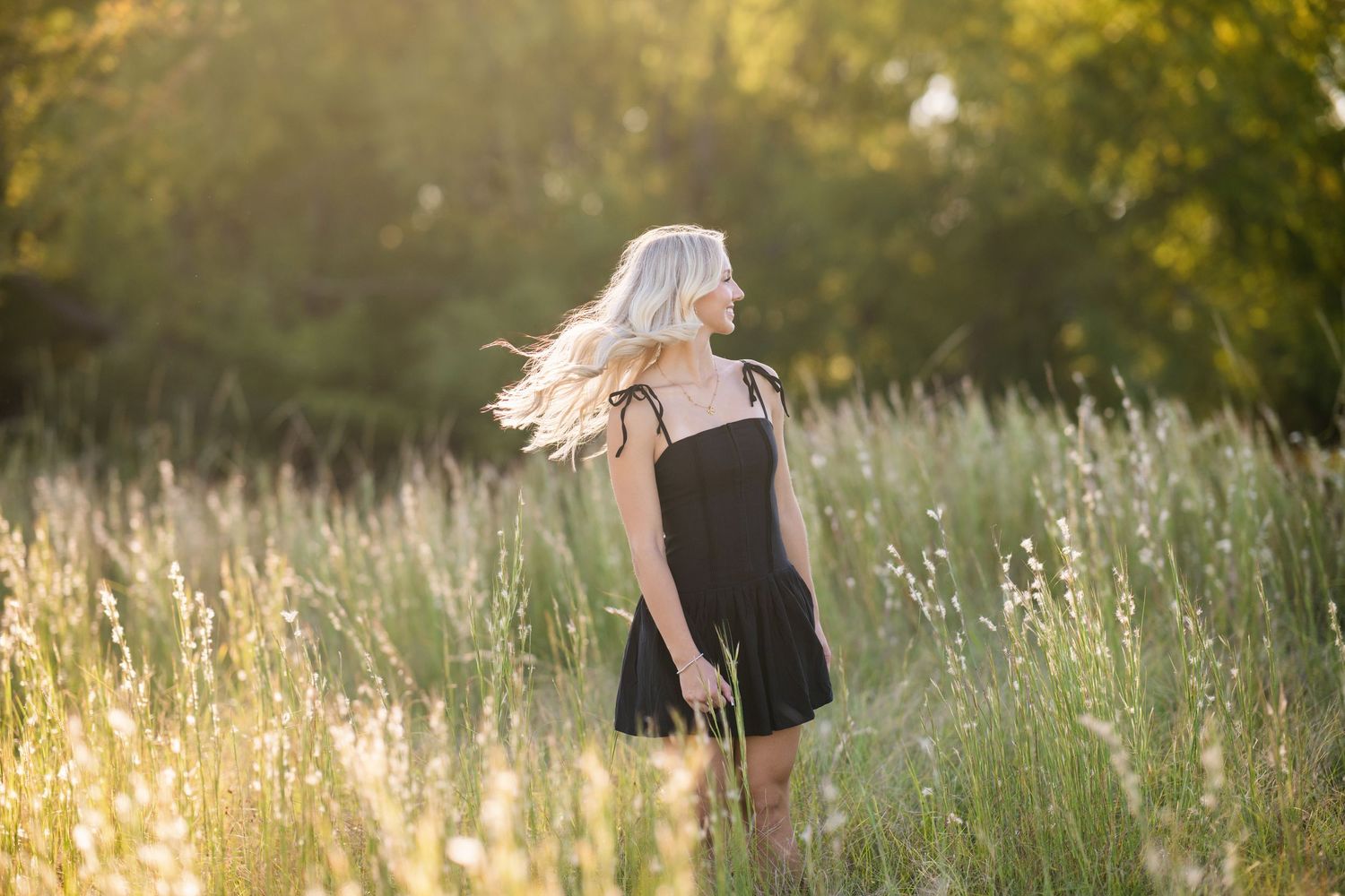 Blonde teen in black sundress stands in golden sunlit field for senior photos.
