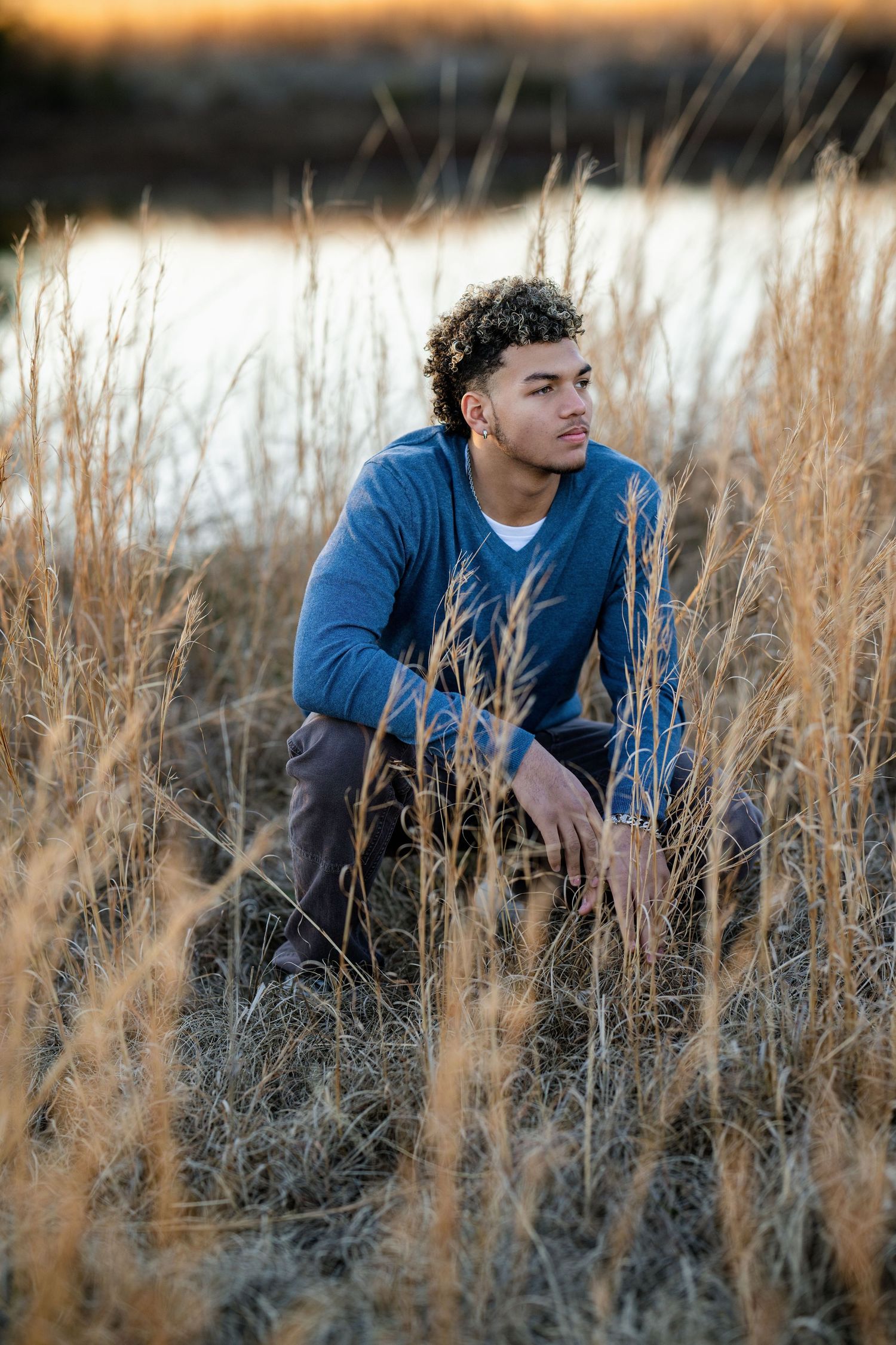 Young man in blue jacket crouching in tall golden grass near water at sunset.