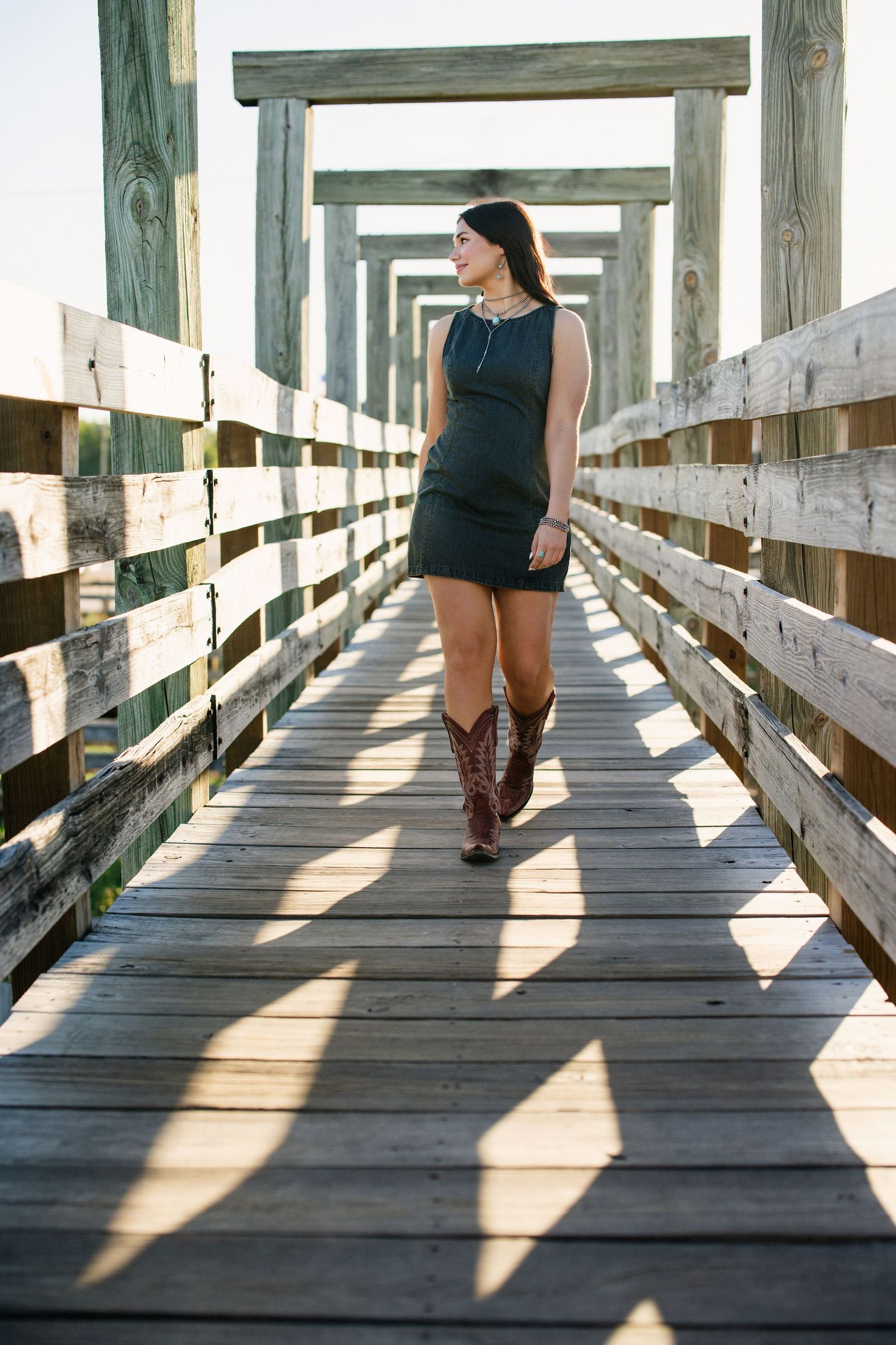 Woman in black mini dress and cowboy boots strolls along sunlit wooden boardwalk.