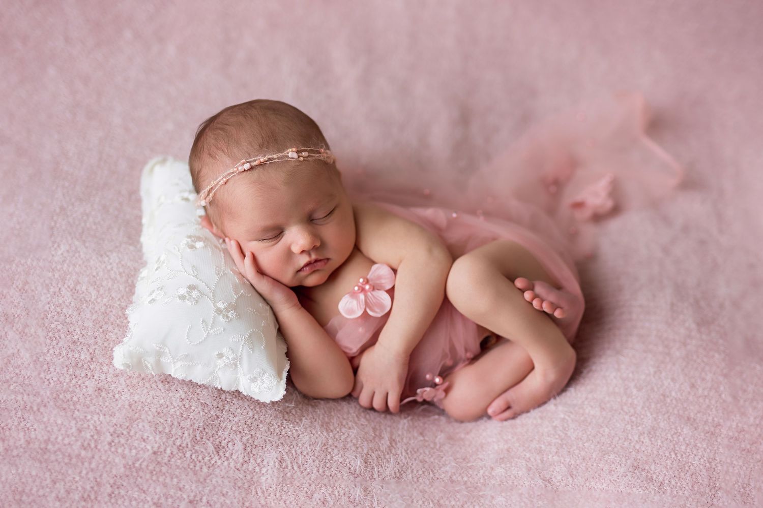 Peaceful newborn baby girl in pink outfit sleeps on white pillow in professional studio photo.