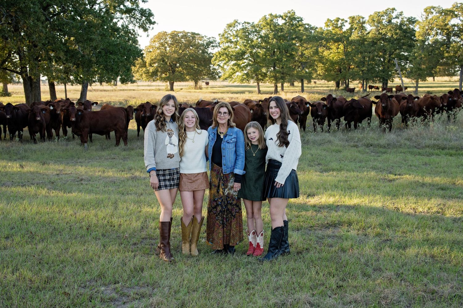 Five girls posing together in a pasture with red cattle grazing under oak trees.