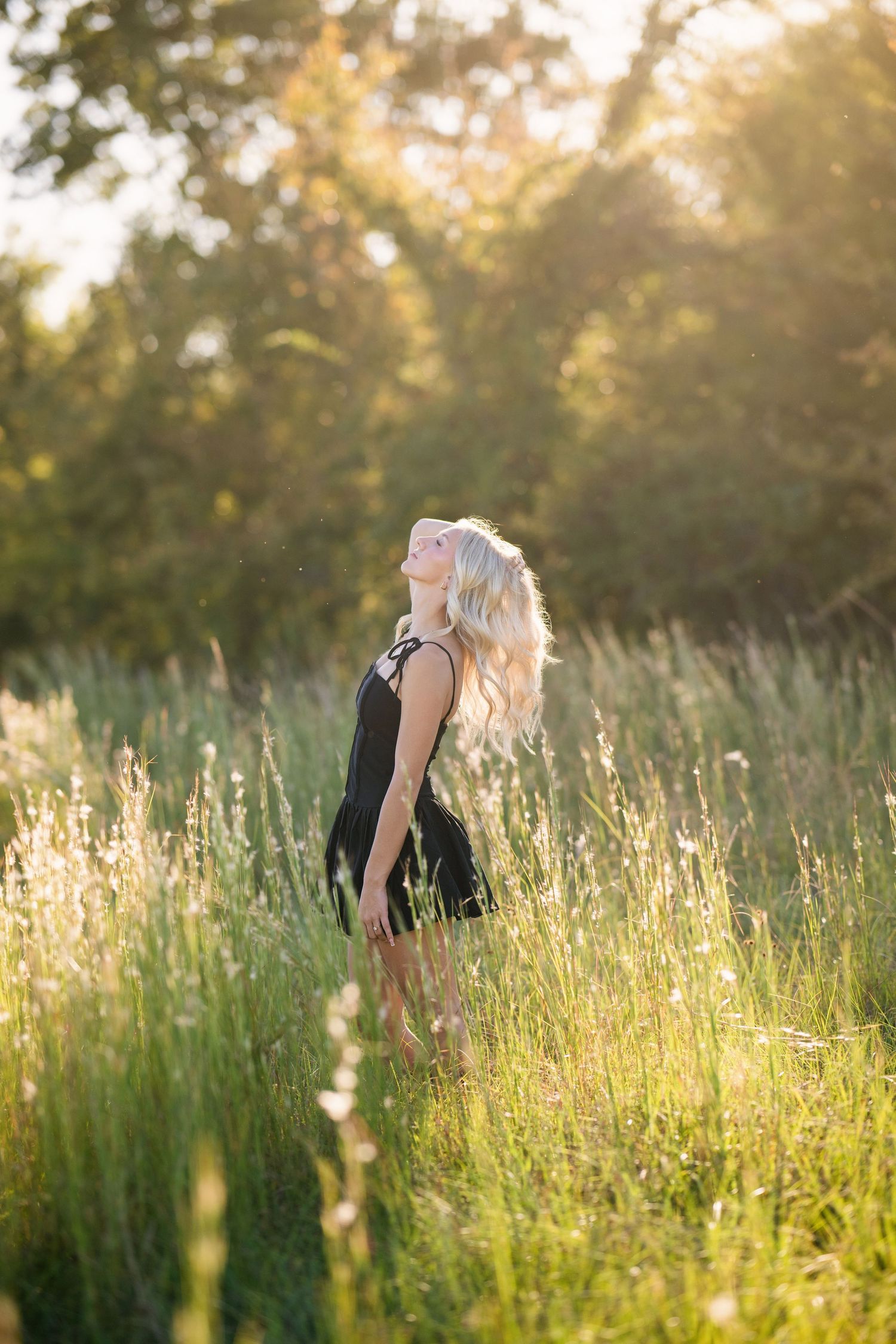 Blonde teen in black dress tilts head back in glowing golden sunlit meadow.