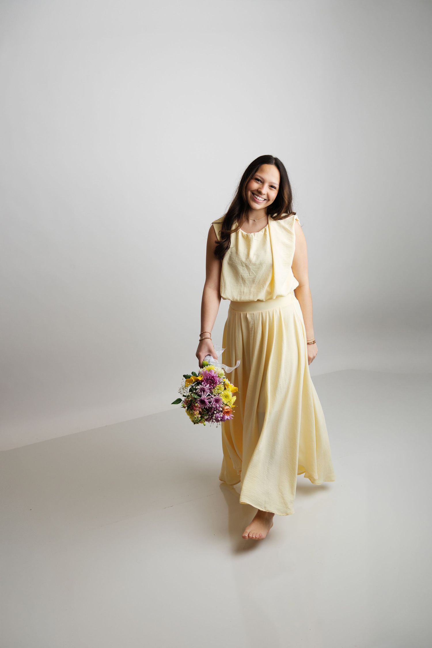 Smiling woman in yellow maxi dress holds colorful flower bouquet in studio.