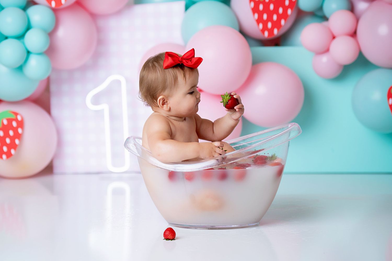 Adorable baby girl in pink bow sitting in milk bath tub with strawberries at first birthday.