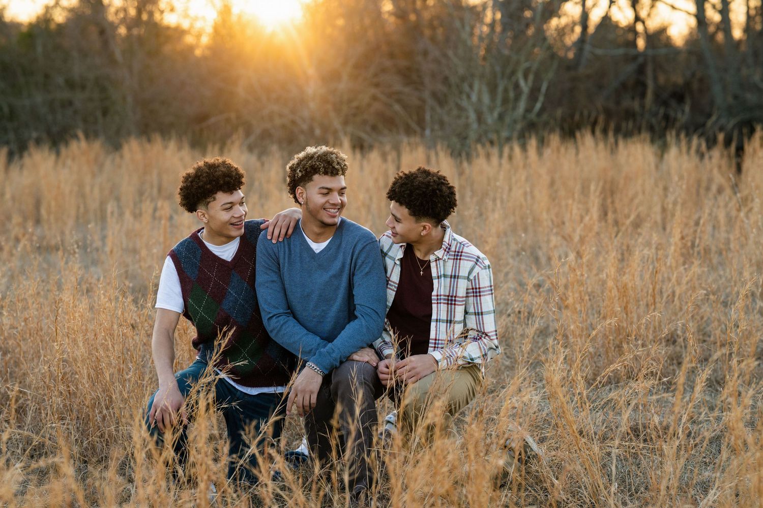 Three teenage brothers sit together smiling in a golden field with warm sunset light glowing behind them.