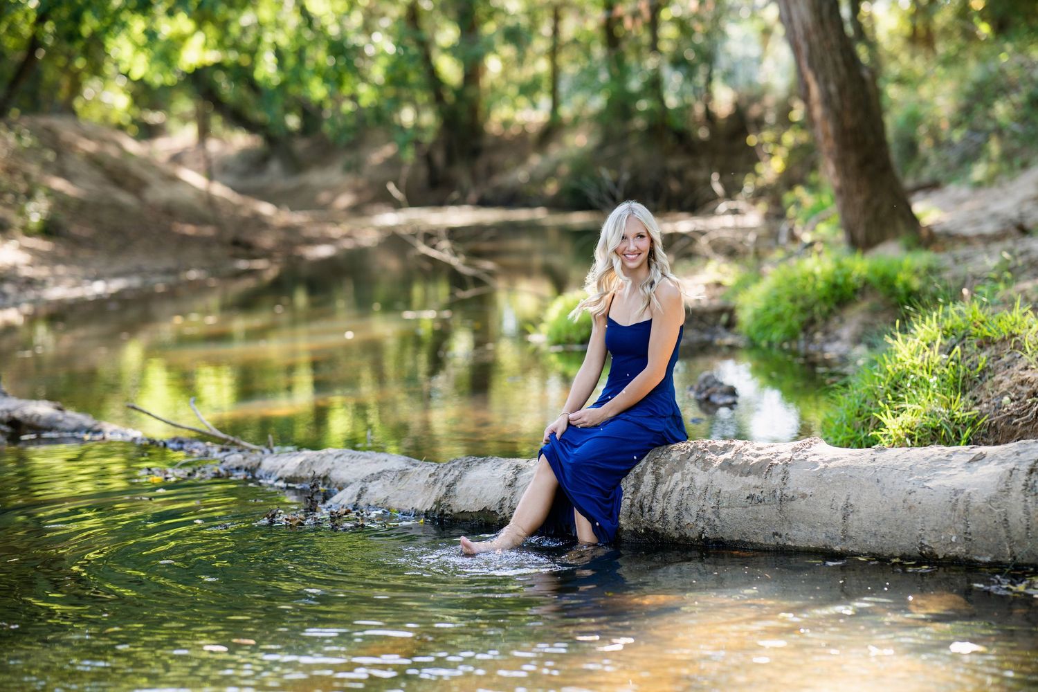 Young blonde woman in a blue dress sitting on a log over a peaceful forest creek, feet in water.