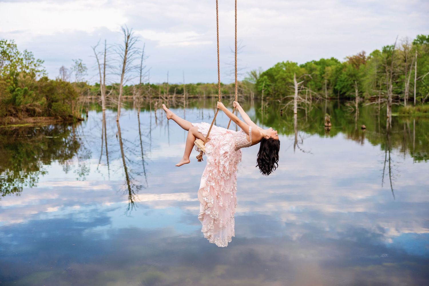 Woman in flowing white dress swinging on rope swing above calm lake with cloudy sky reflection.