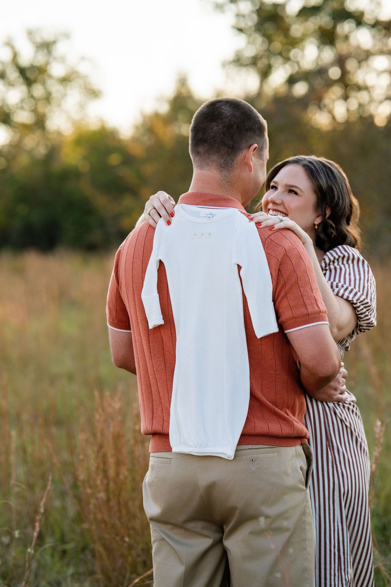 Couple embraces in golden field at sunset while holding tiny white baby onesie announcement.