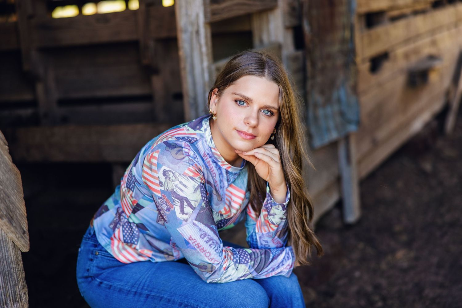 Teenage girl in patterned sweater sitting in rustic barn setting, posing for senior portrait.