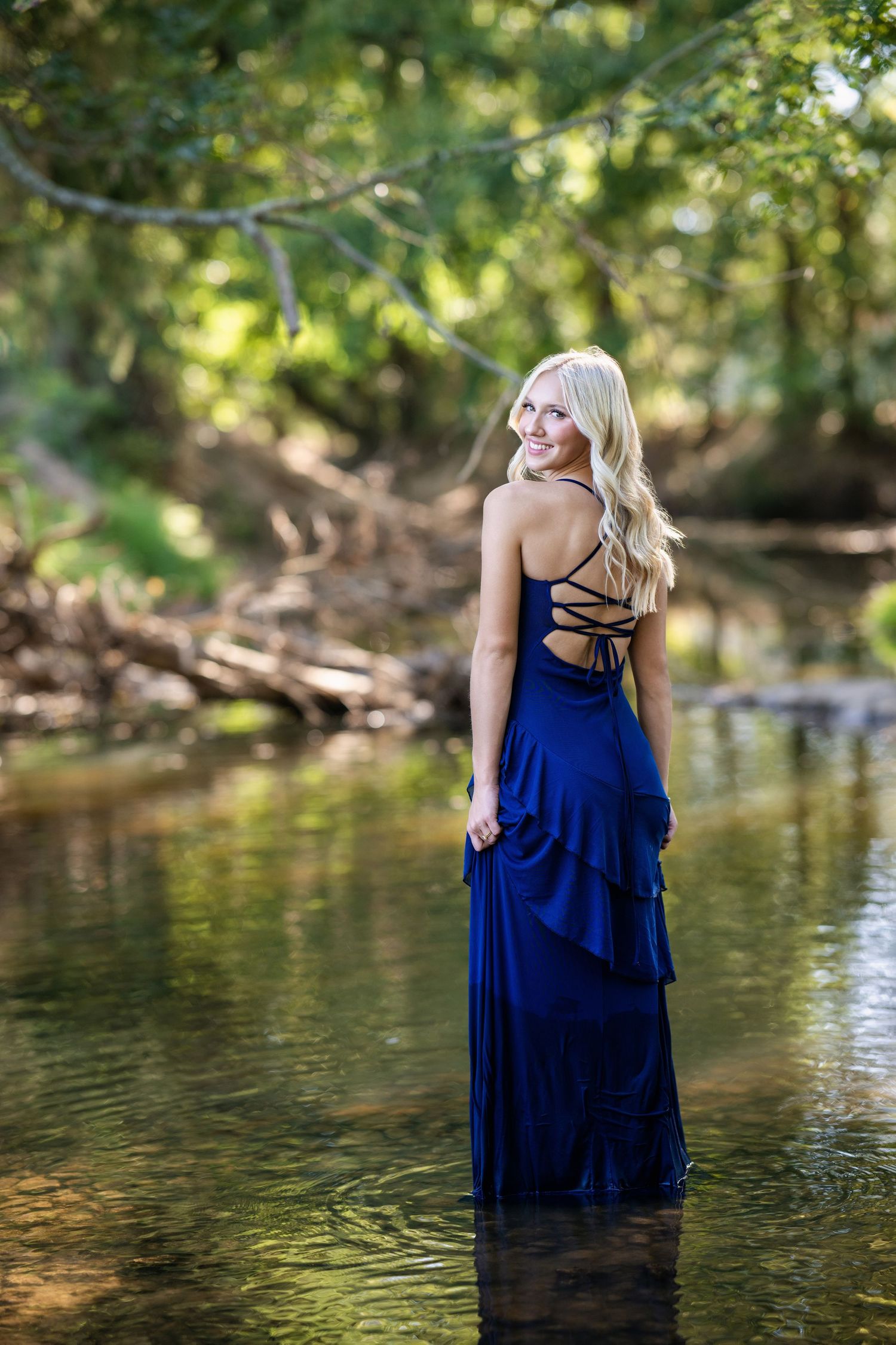 Blonde woman in a navy blue backless gown standing in a serene forest creek, smiling over shoulder.