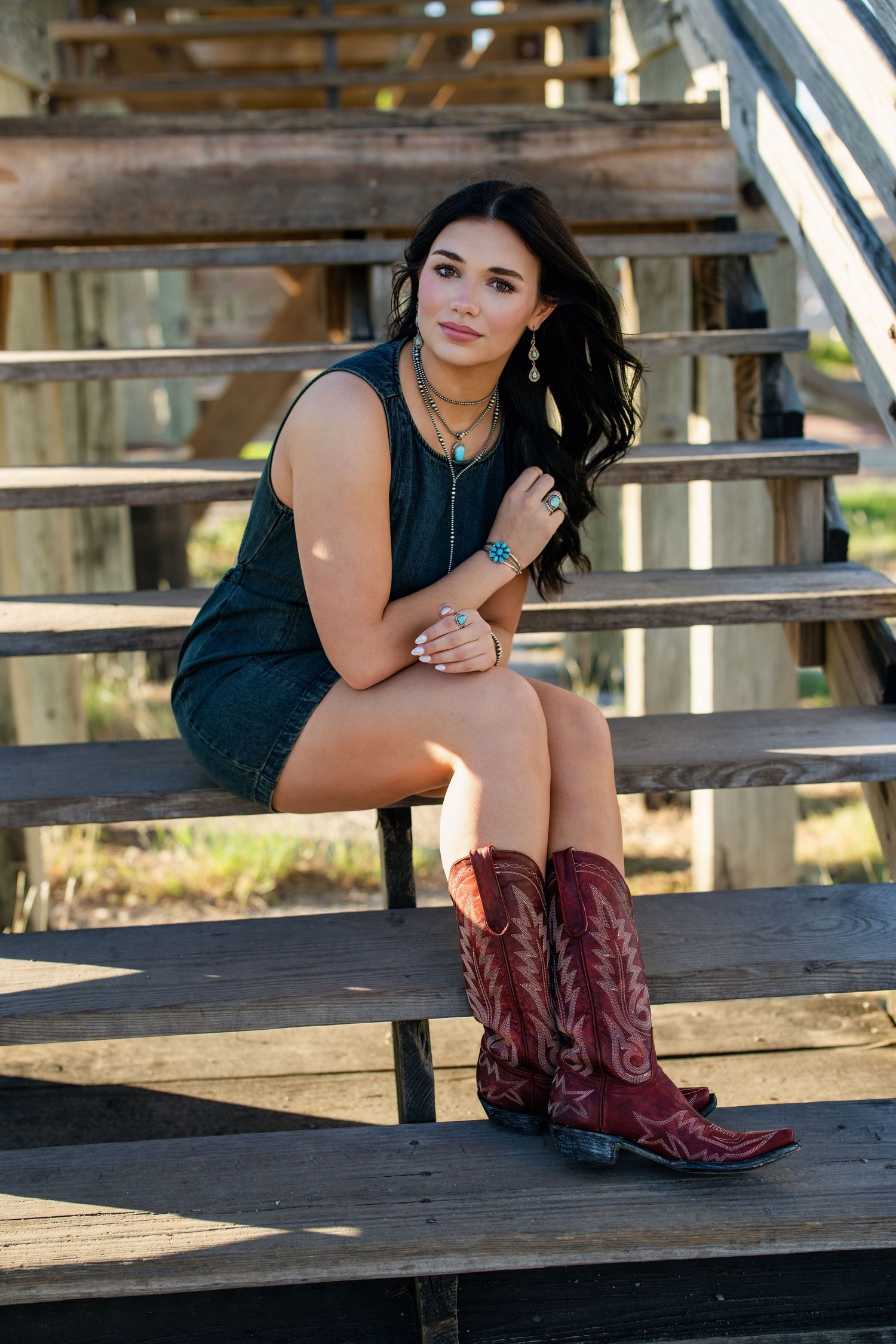 Brunette girl in dark green mini dress and red cowboy boots sitting on wooden bleachers outdoors.