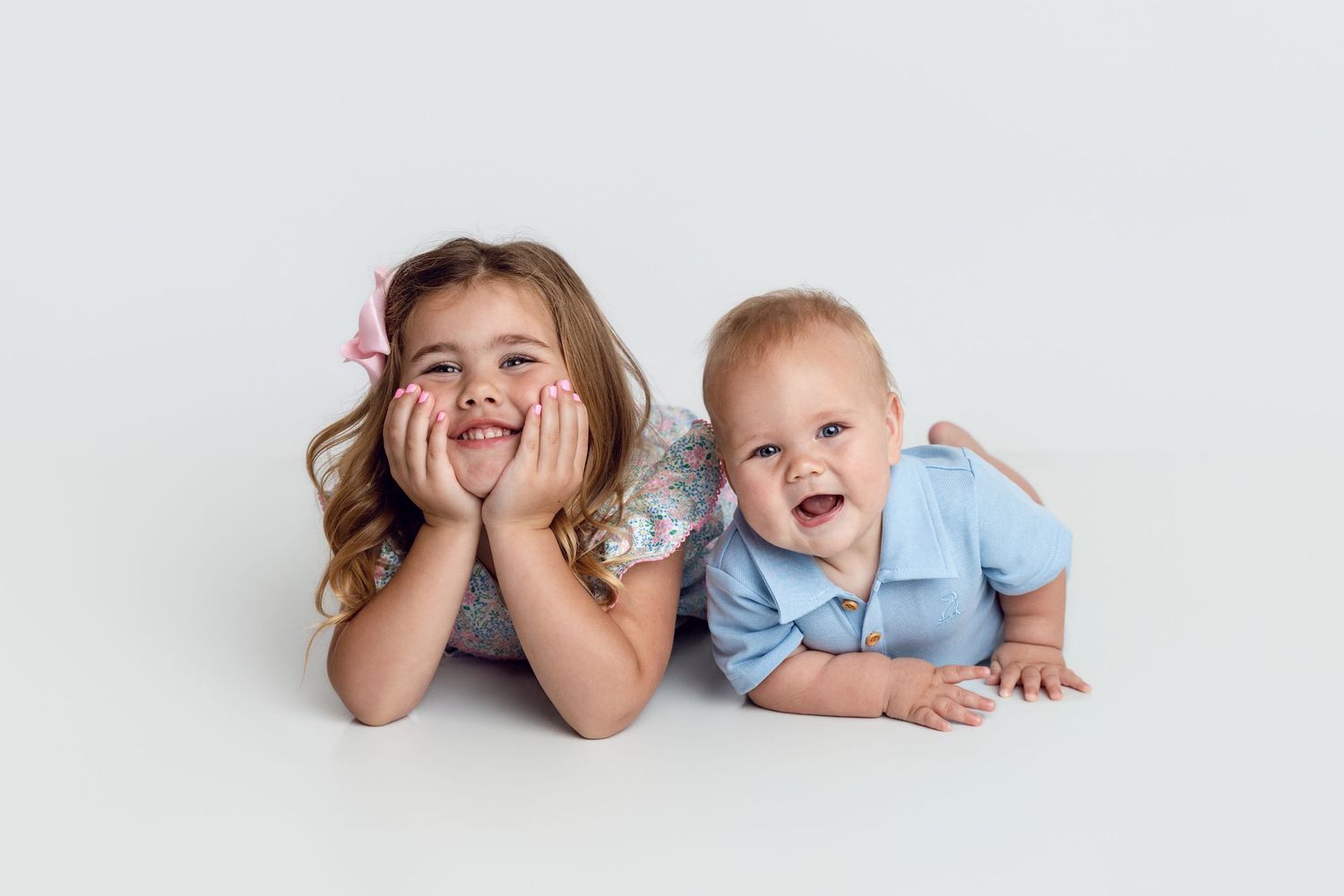 A smiling young girl and a laughing baby boy lie side by side on a white studio floor.