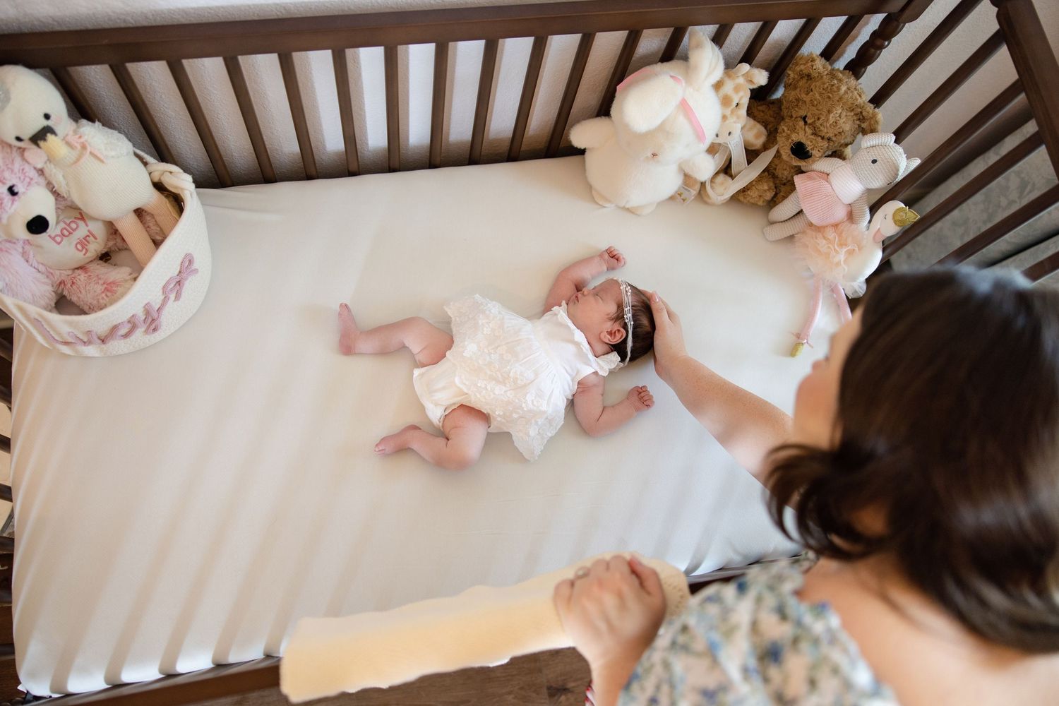 Mother gently tending to newborn baby lying in wooden crib with stuffed animals.