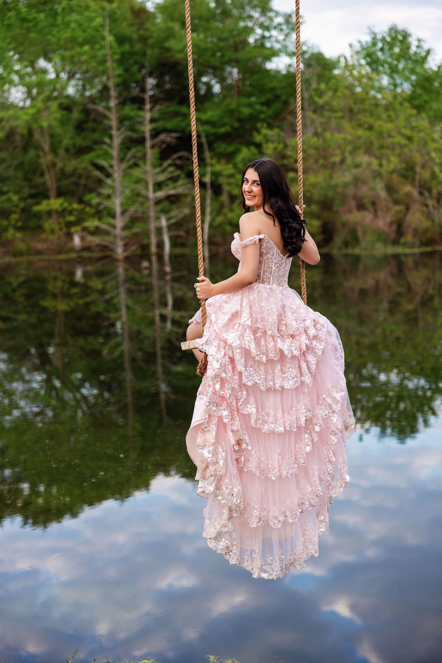 Woman in pink ruffled ballgown sits on rope swing over reflective lake.