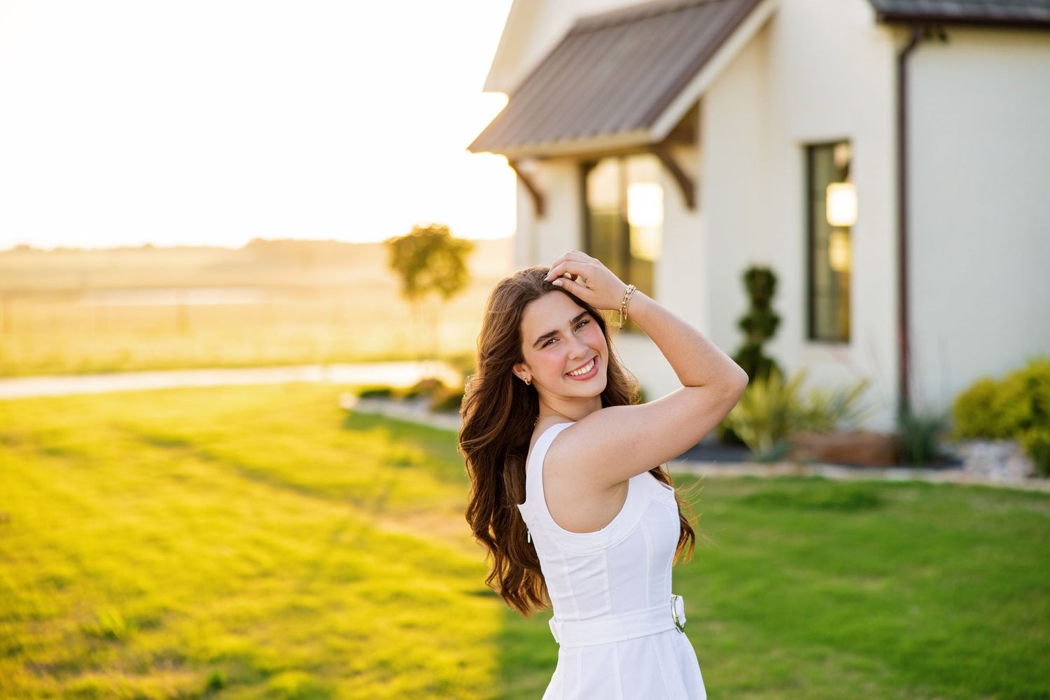 Smiling young woman in white sundress poses outdoors near a farmhouse at golden hour sunset.