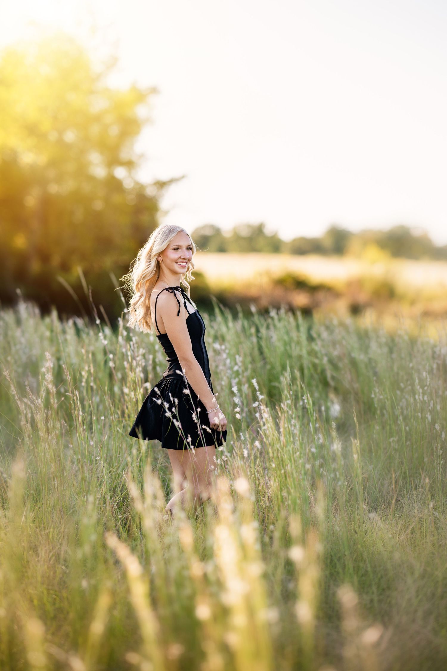 Smiling blonde girl in black dress poses in tall grass field during golden hour.