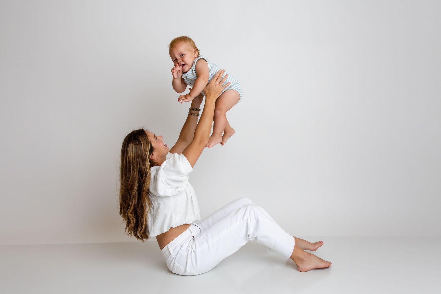 A mother sits on a white floor lifting her smiling baby up in the air during a studio photo session.