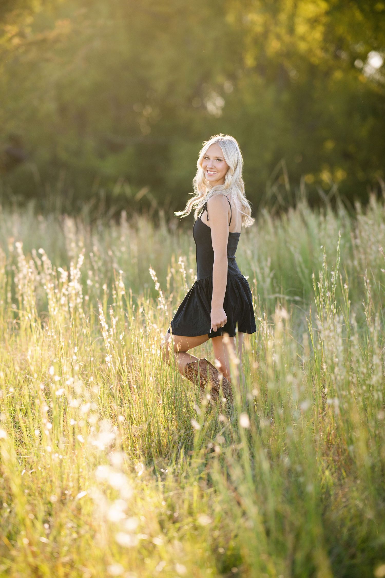 Laughing blonde senior girl in black dress poses in wildflower field at sunset.