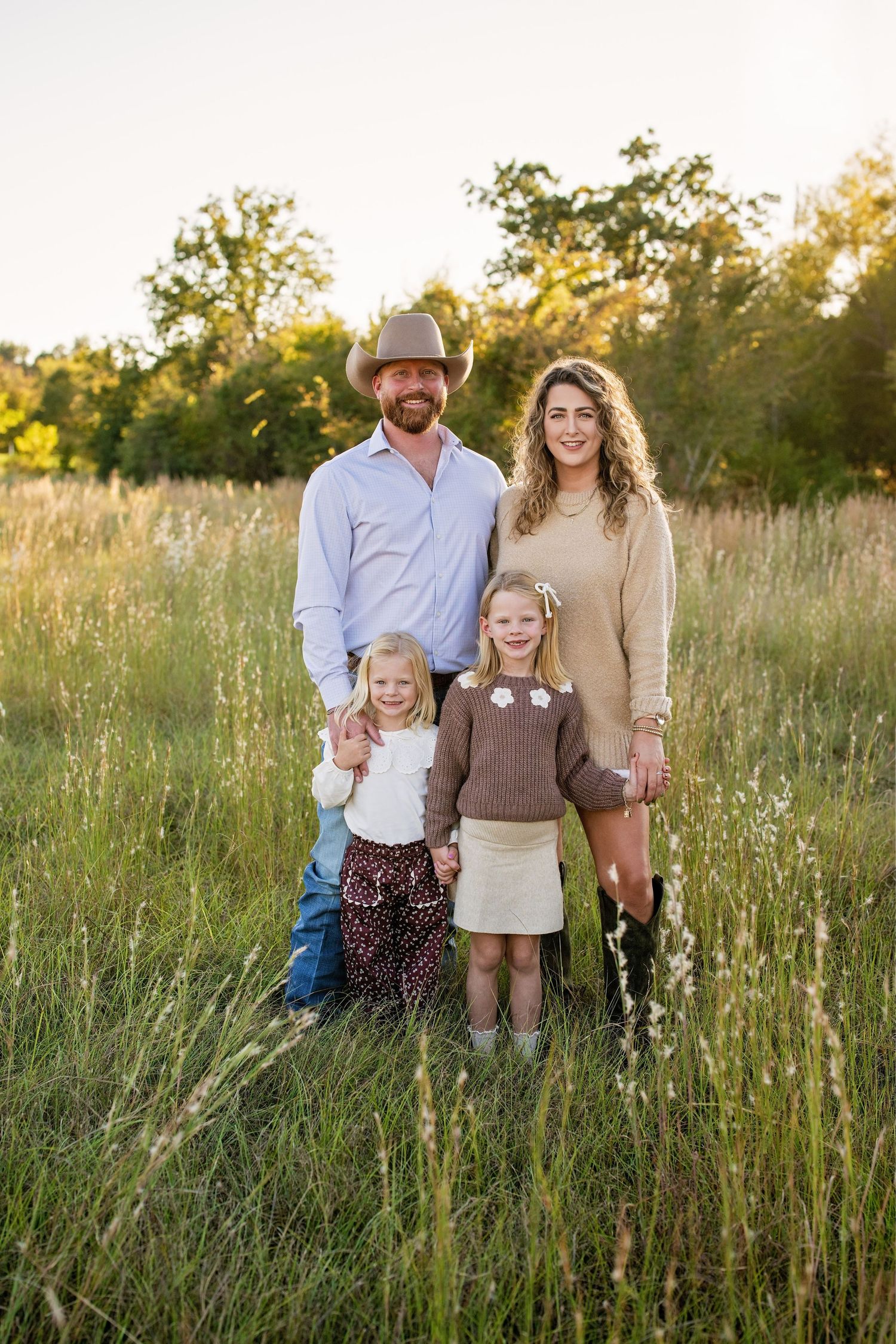 Happy family of four posing in green meadow at golden hour with trees behind them.