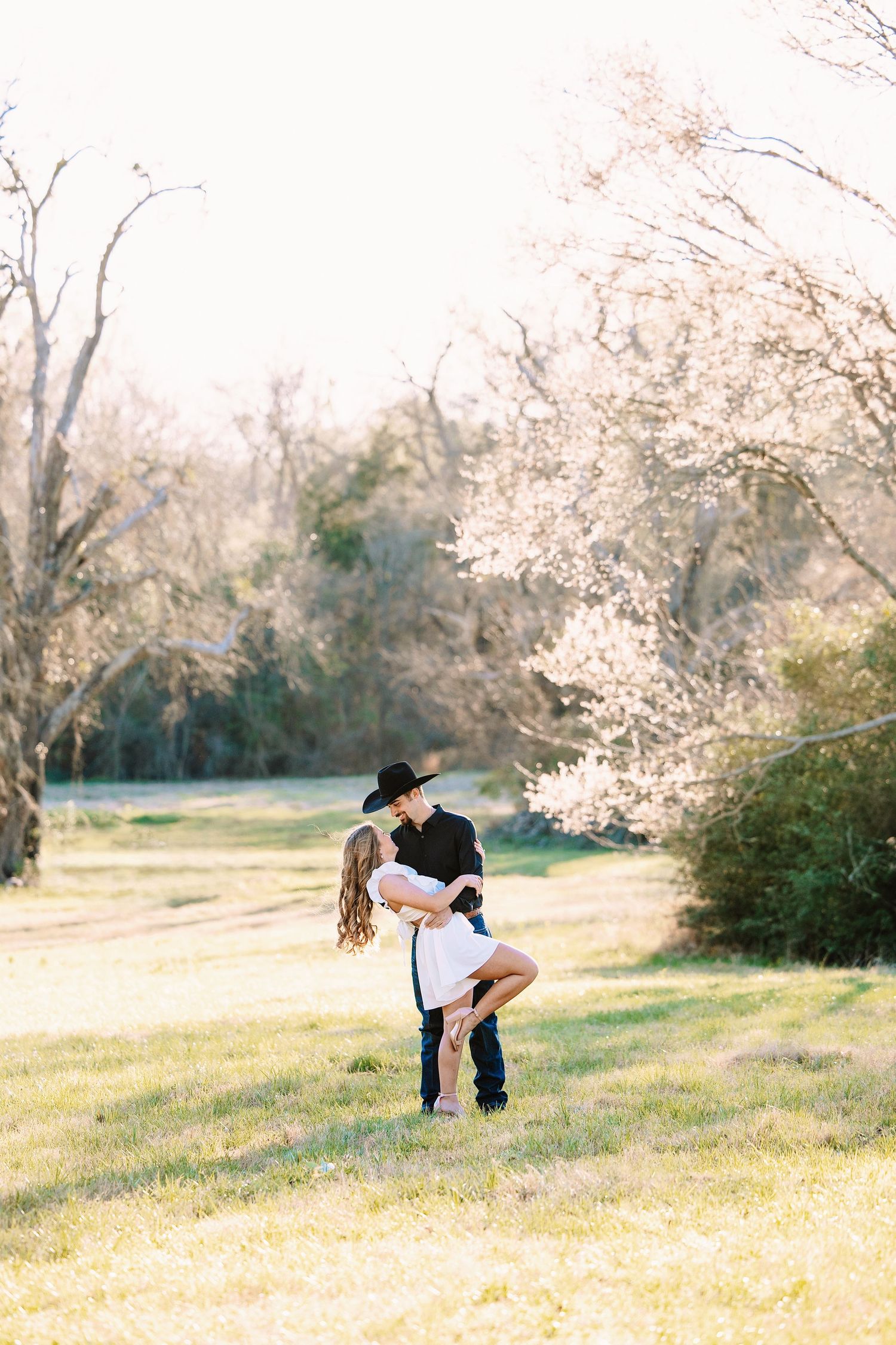Romantic couple embracing in sunlit field with blooming trees, man wearing black cowboy hat.