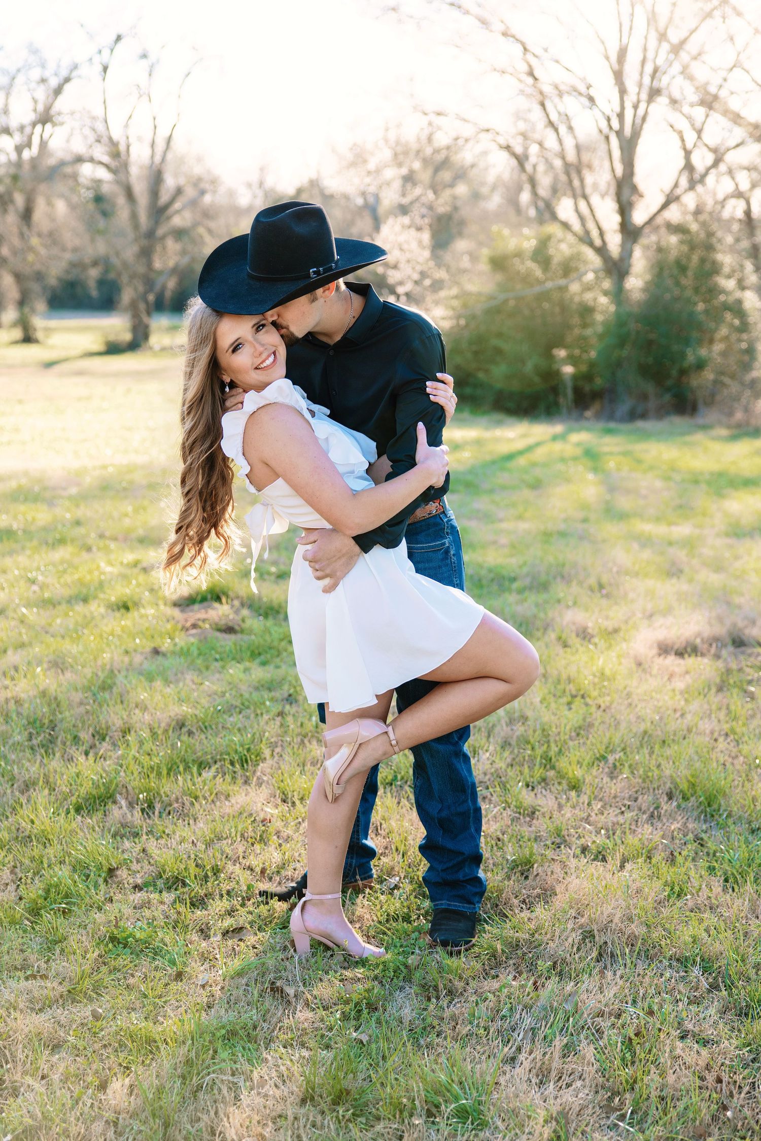 Couple embracing in golden hour light, man in black cowboy hat lifting woman in white dress.