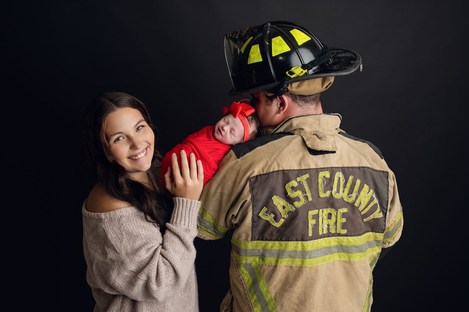 Smiling mother holds newborn baby beside proud father in East County Fire firefighter uniform.