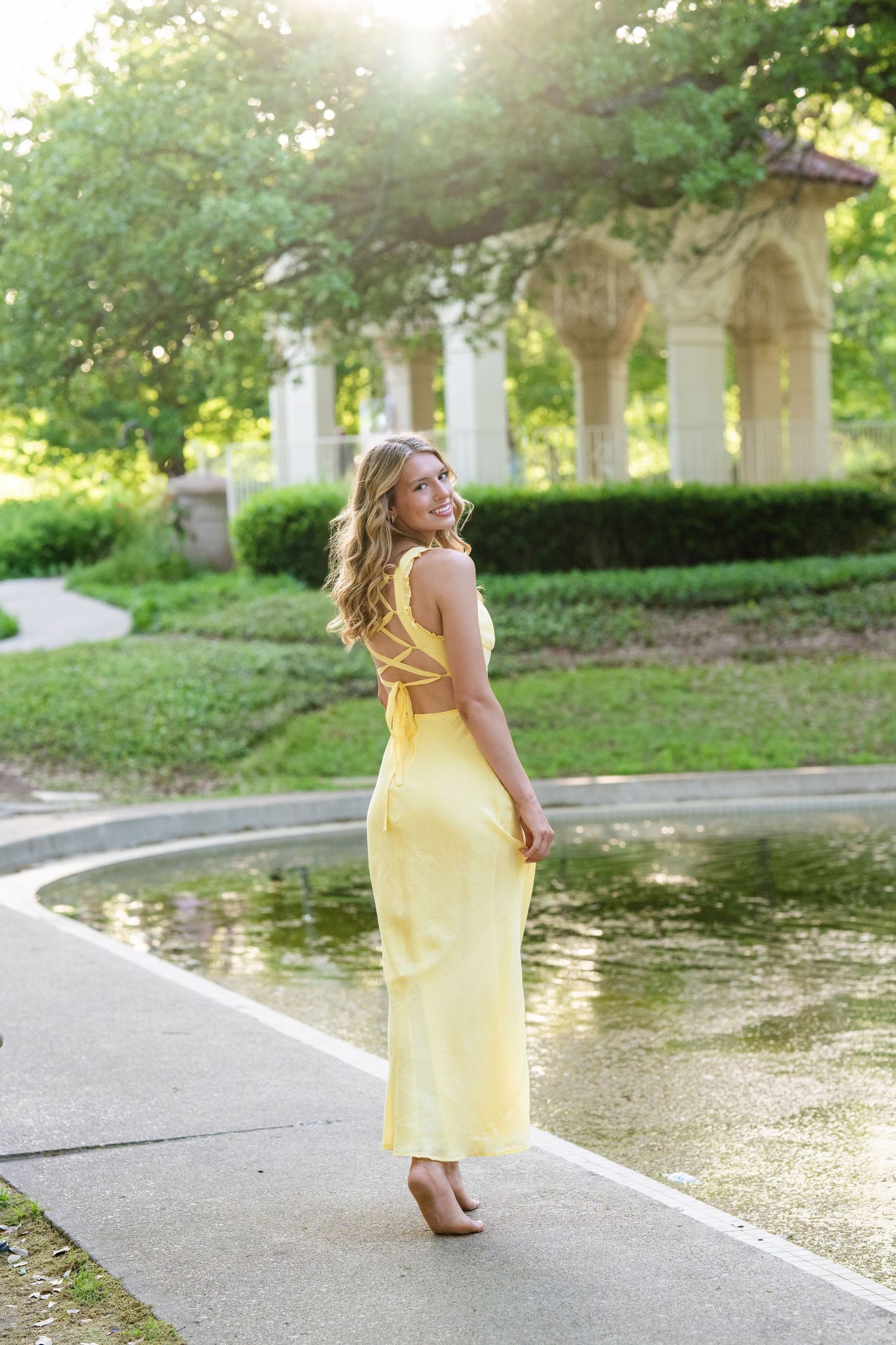 Blonde girl in yellow backless maxi dress posing near fountain with white gazebo for senior photos.