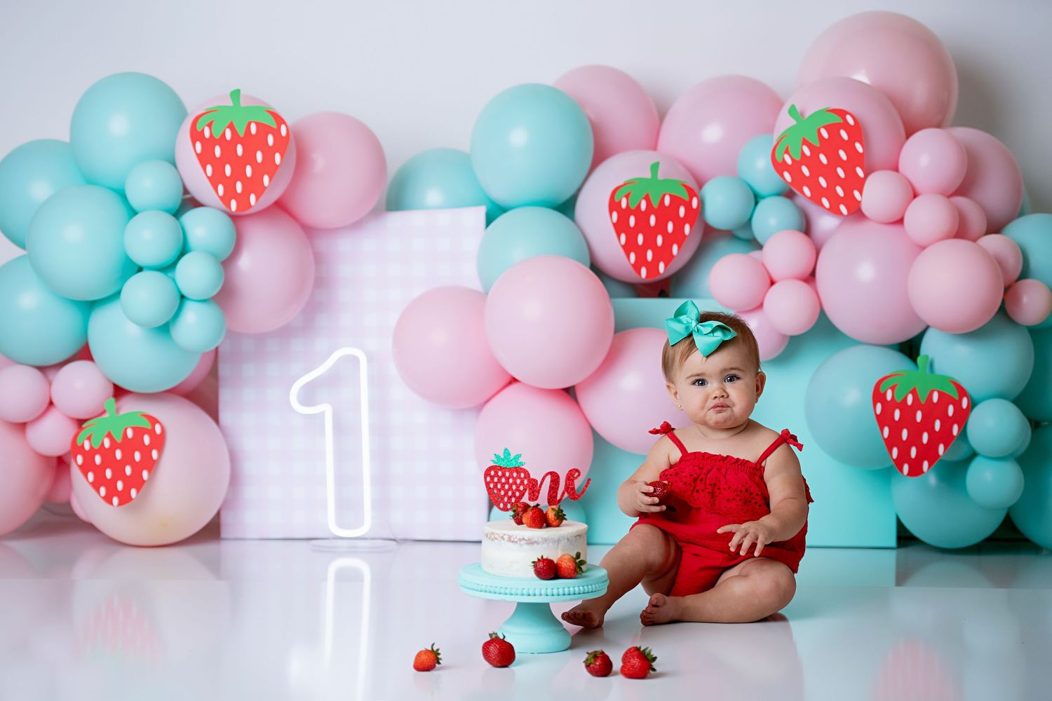 Baby girl in red outfit celebrates first birthday with a strawberry themed cake smash and balloon backdrop.
