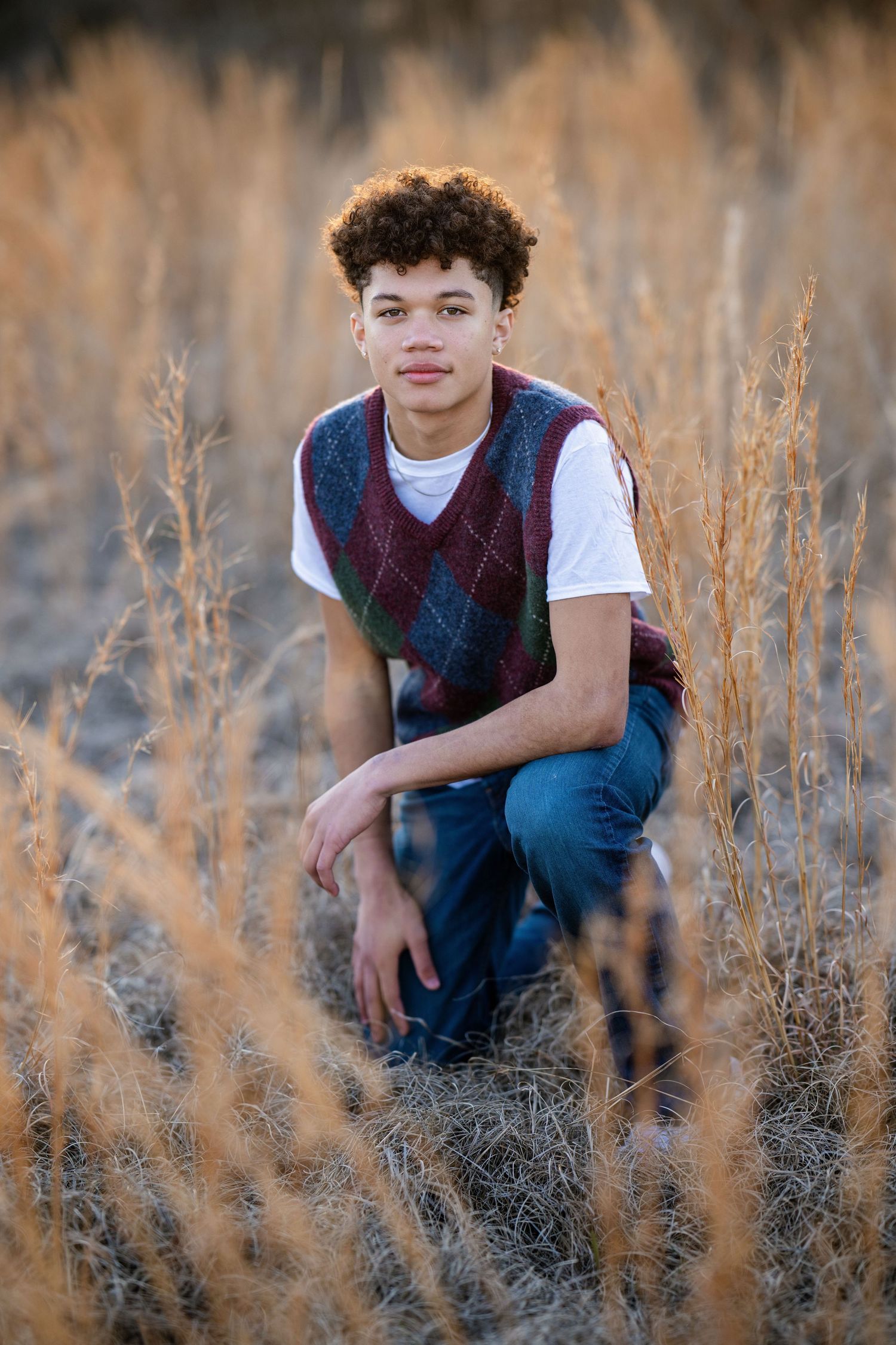 Teen boy in argyle vest and jeans crouches in dry golden grass for senior portrait.