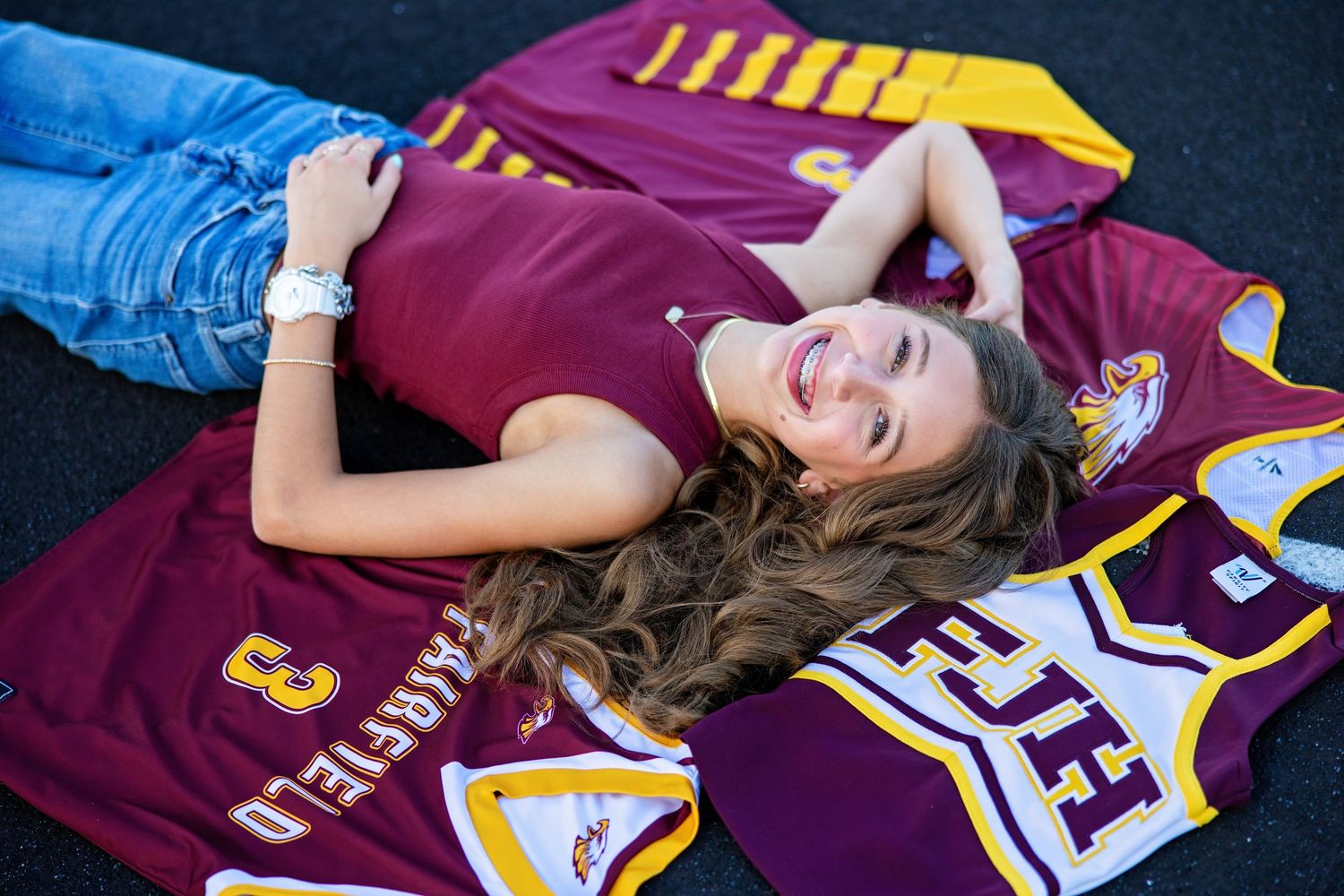 Smiling teen girl lying on sports jerseys on a track, wearing jeans and maroon top.