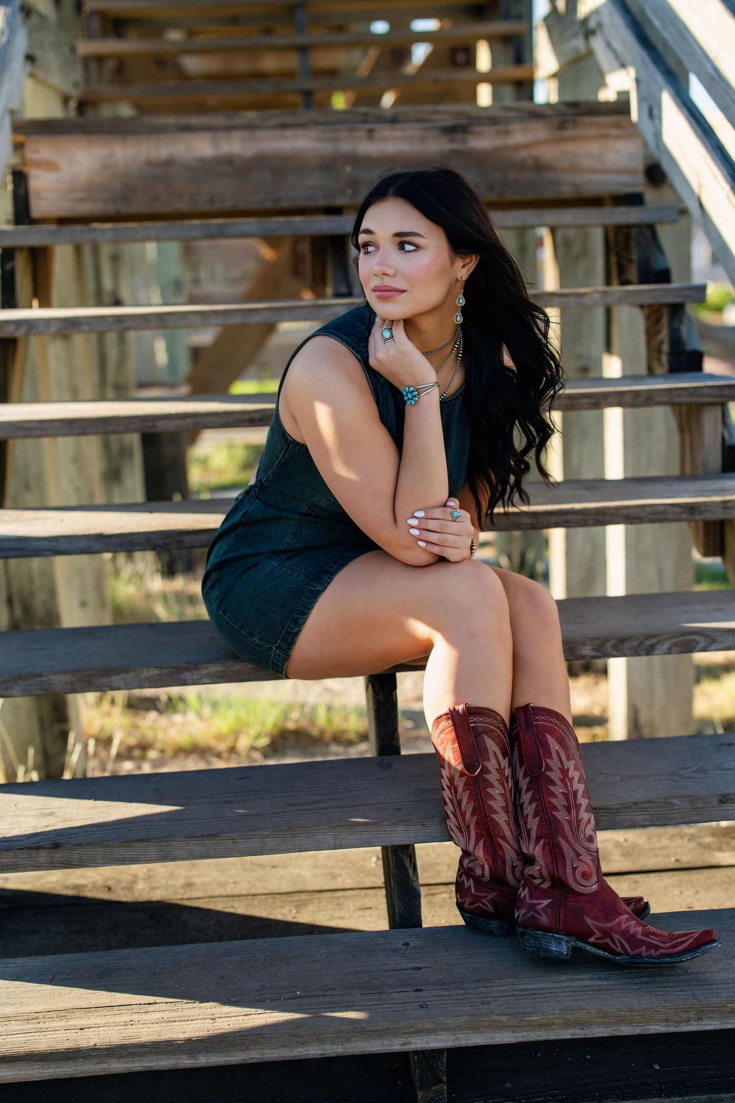 Brunette woman in dark mini dress and red cowboy boots sits on wooden bleachers.