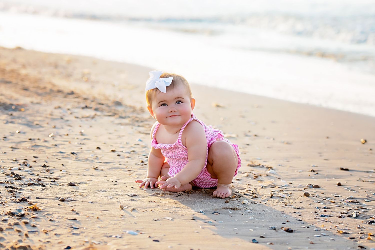 Cute baby girl in pink ruffled swimsuit crawling on sandy beach near ocean waves.