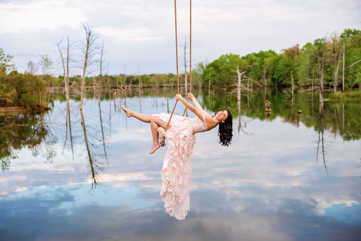 Woman in white lace dress swinging on rope swing over reflective lake surrounded by trees.
