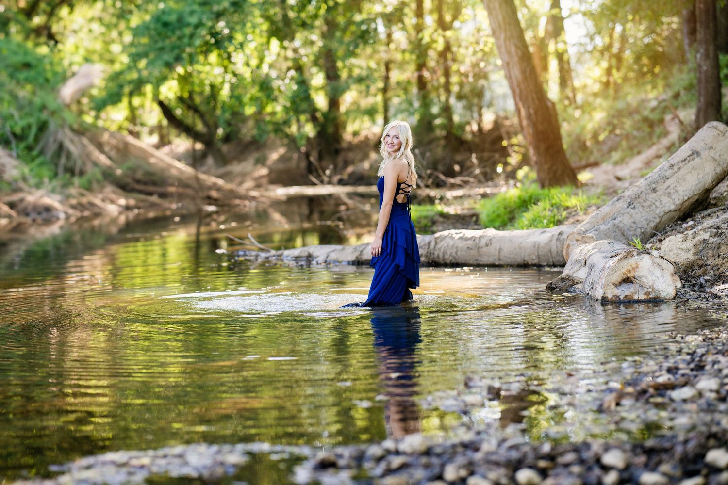 Blonde woman in a flowing navy blue dress wading through a sunlit forest creek.