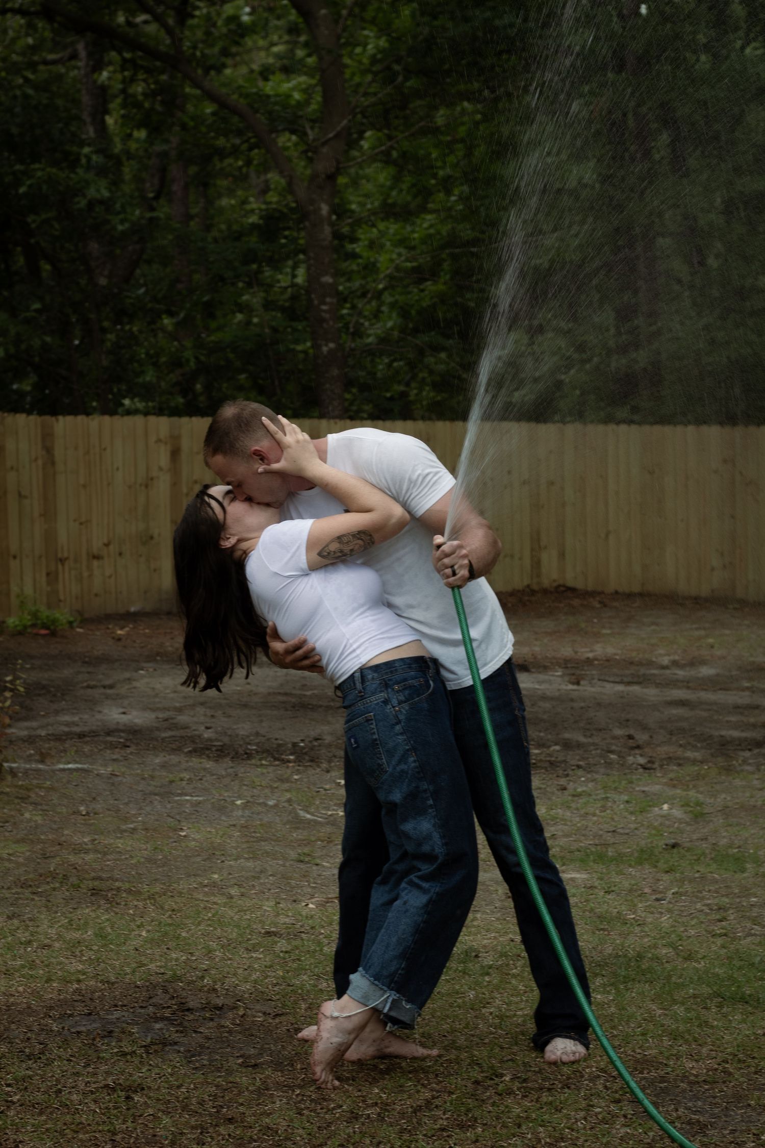 Parents sharing a sweet moment together spraying garden hose while kissing during an outdoor family session in Raeford, NC.