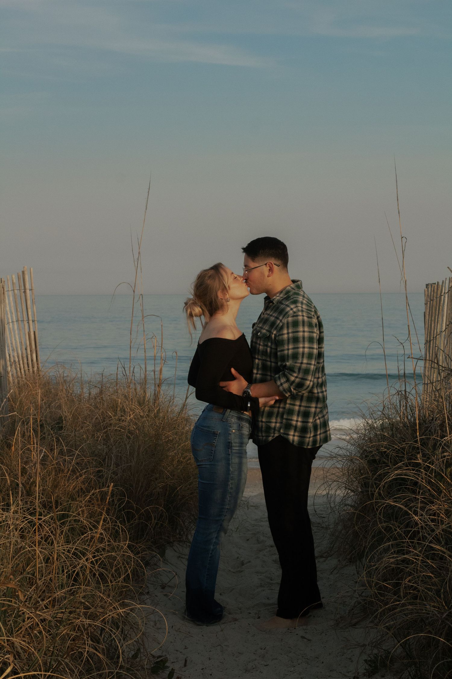 Couple kissing beside a wooden beach fence at sunset during their Wrightsville Beach engagement session.