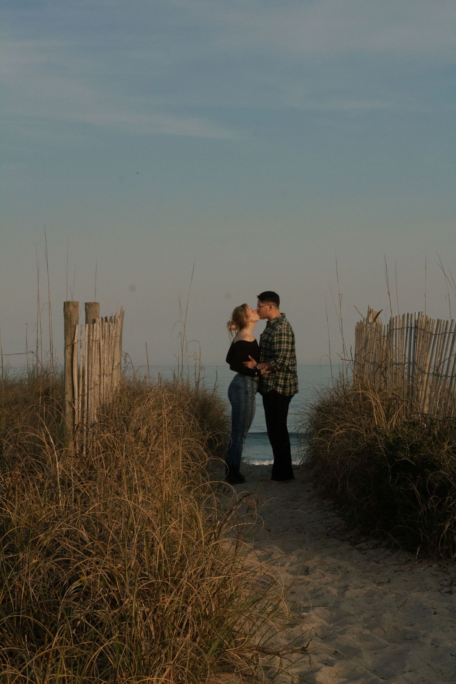 Couple kissing beside a wooden beach fence at sunset during their Wrightsville Beach engagement session.