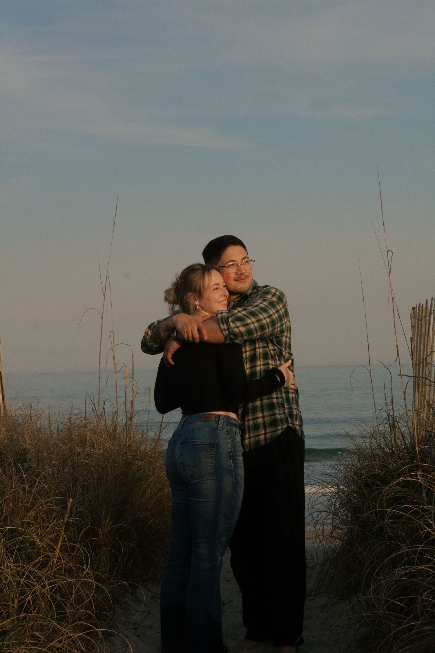 Couple embracing beside a wooden beach fence at sunset during their Wrightsville Beach engagement session.
